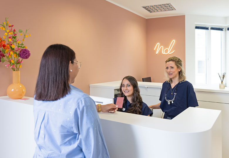 Zahnärztin und Rezeptionistin im Gespräch mit einer Patientin am Empfangstresen.