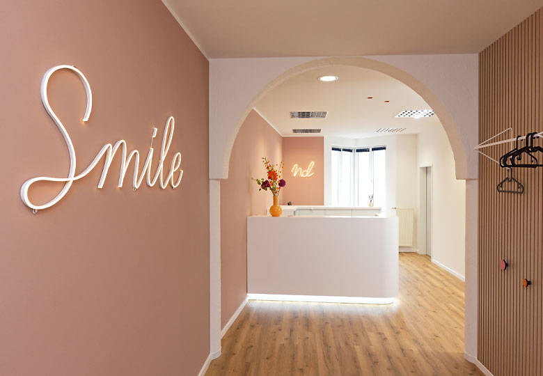 Modern gestalteter Empfangsbereich unserer Zahnarztpraxis mit 'Smile' Leuchtschrift und hellem Tresen in pastellfarbener Umgebung.
